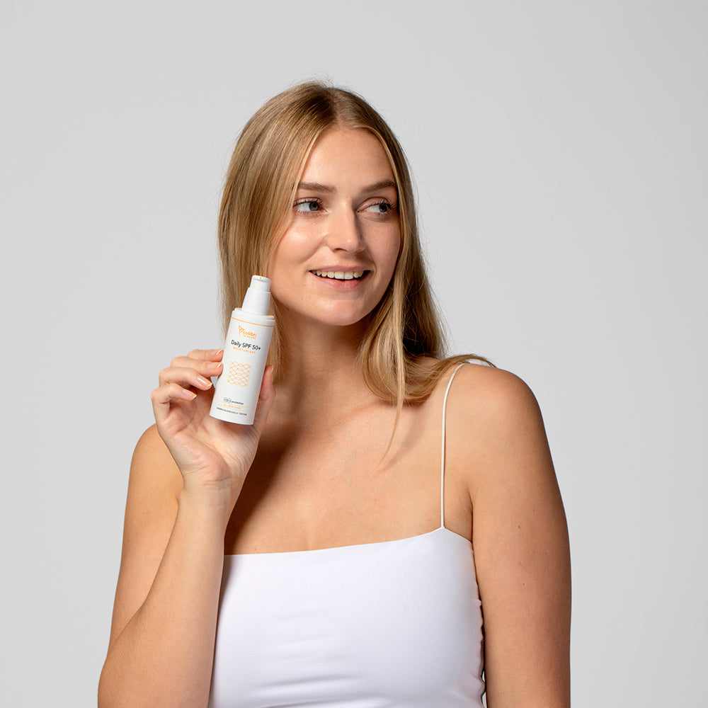 Eine Frau im weißen Top hält eine Flasche mit Hautpflegeprodukt und lächelt.